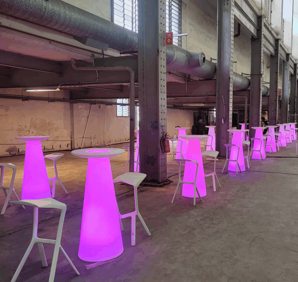 Rows of tall tables glowing with purple light are paired with white bar stools in an industrial warehouse space with exposed ducts and pipes.