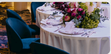 A round table with a white tablecloth set for a meal, featuring floral centerpieces and blue chairs arranged around it.